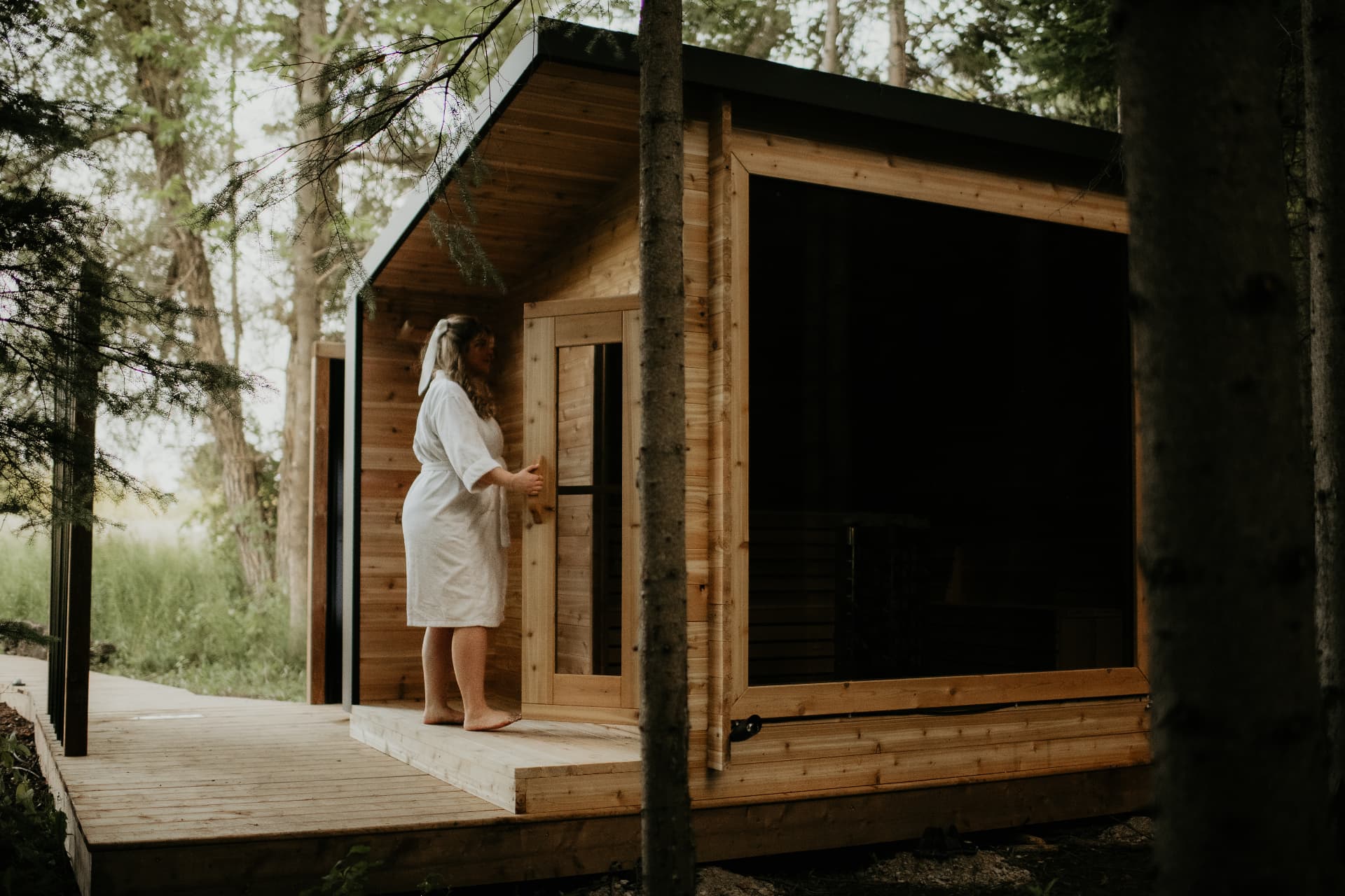 Woman in a white robe entering a cedar prefabricated sauna in a wooded setting